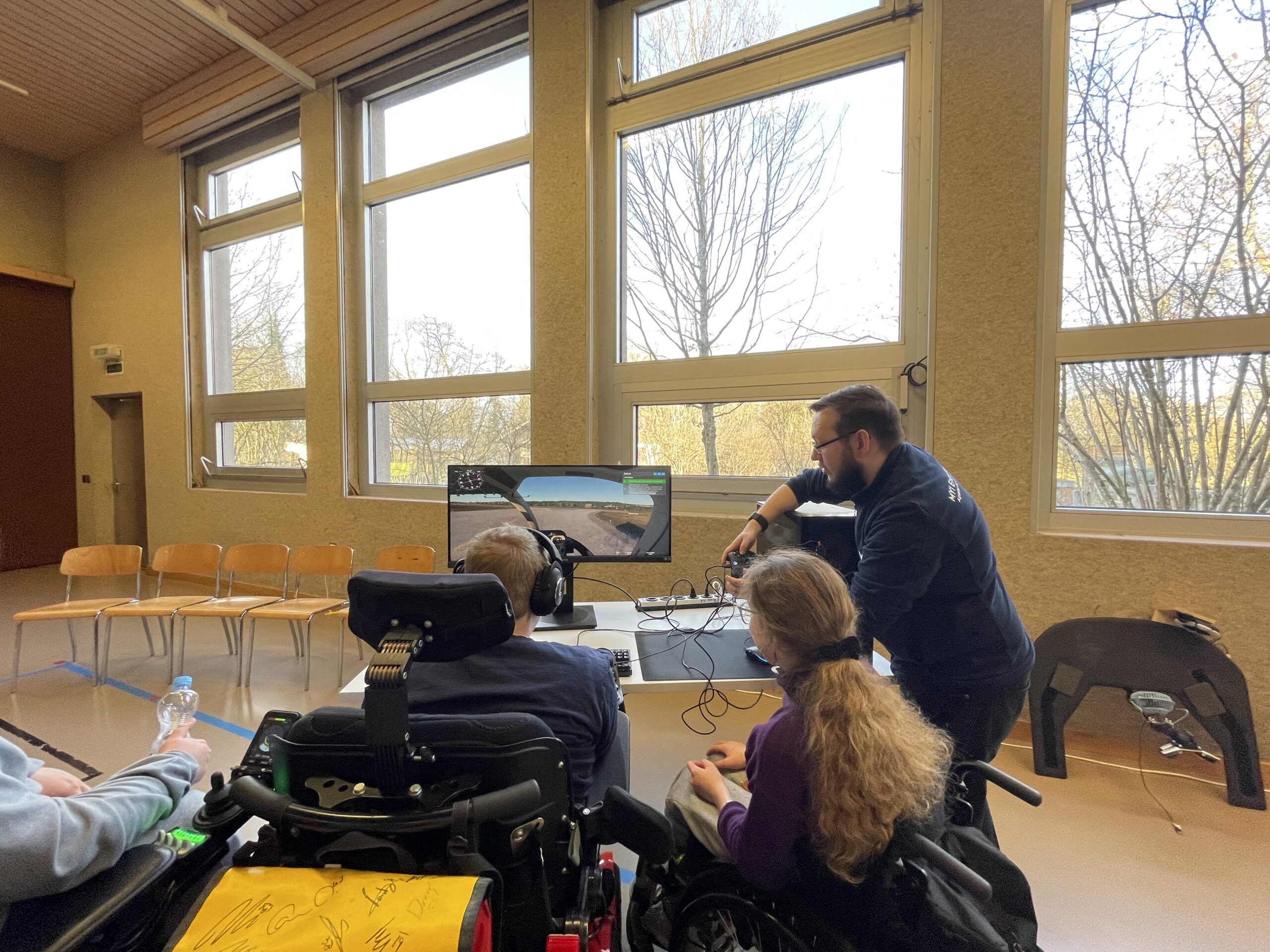 Zwei Kinder mit Rollstuhl spielen ein Game in einer Turnhalle. Ein Betreuer unterstützt sie.