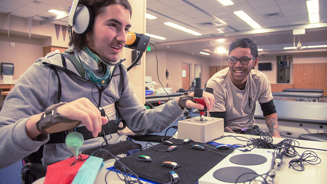 Ein Gamer spielt mit einem Xbox Adaptive Controller. Eine zweite Person schaut zu und lacht.